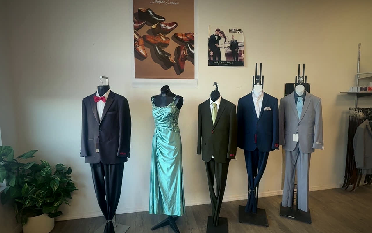 Four mannequins in a dark suit with a red bow tie, an olive suit, a navy suit and a light grey suit, beside a fifth in a teal satin gown