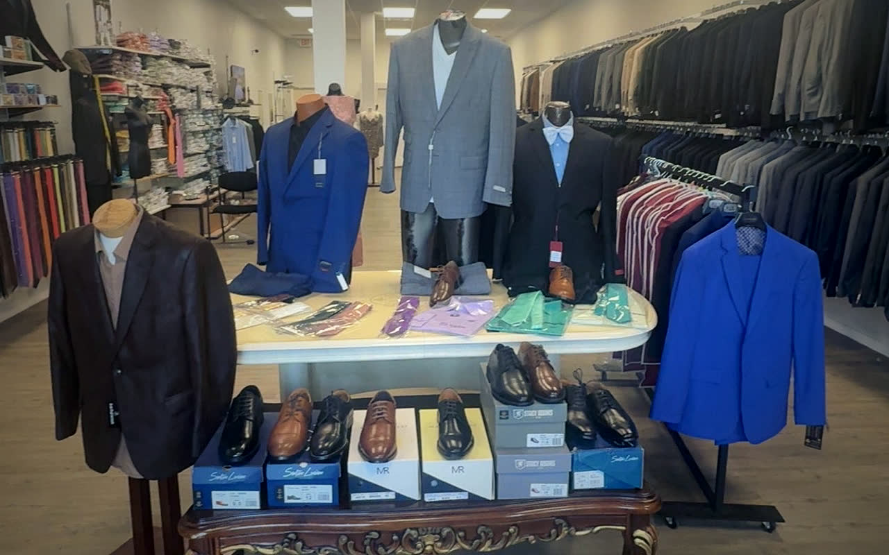 Front of the store: dress shoes on a carved wooden table, mannequins in a brown leather jacket, a royal blue suit, a grey check suit and a black tuxedo with a white bow tie, and long racks of suits behind