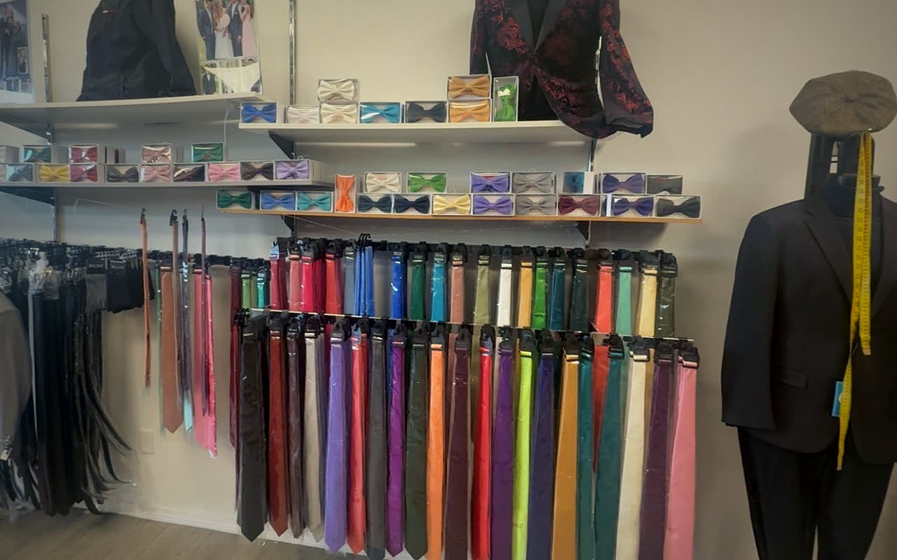 A wall of solid-color ties hanging in two rows, boxed bow ties on the shelves above and belts to the left